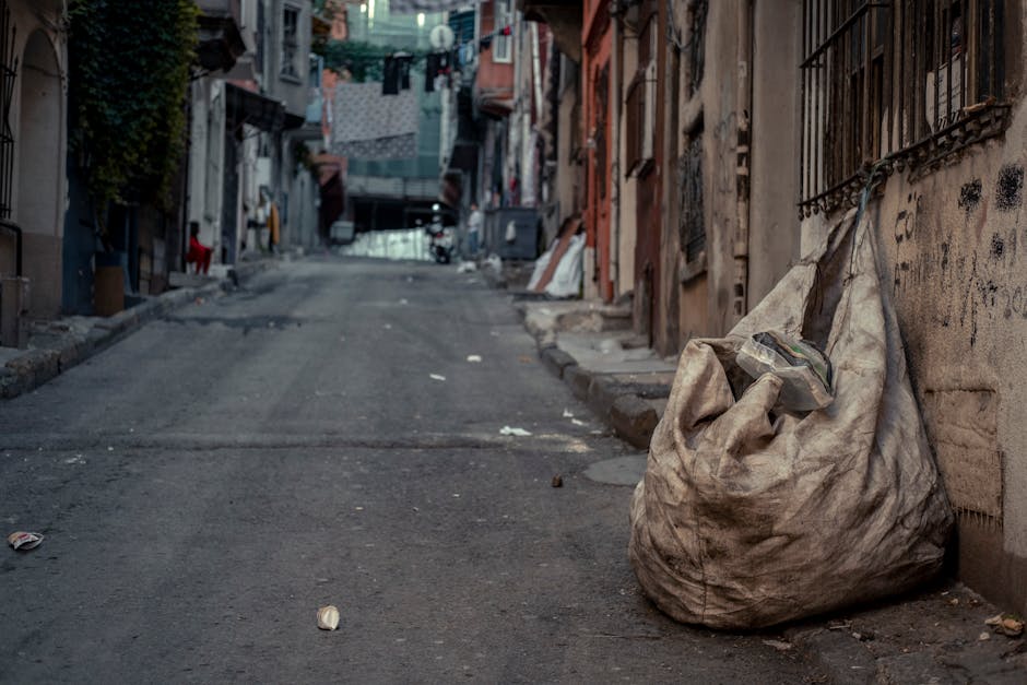 A pile of mixed cardboard boxes, some flattened and others partially intact, along with plastic bags and packaging materials, is located outdoors next to a brick wall and a rough stone base. The cardb